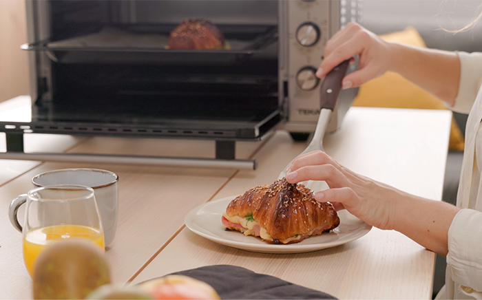 mujer dejando un croissant en una plato con un horno portatil de fondo y un vaso de zumo de naranja