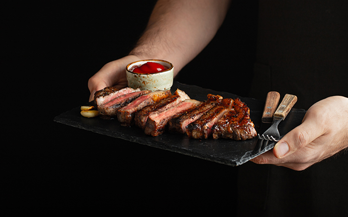 Hombre sujetando una pizarra con un filete partido y cubiertos hecho en barbacoa o en horno SteakMaster