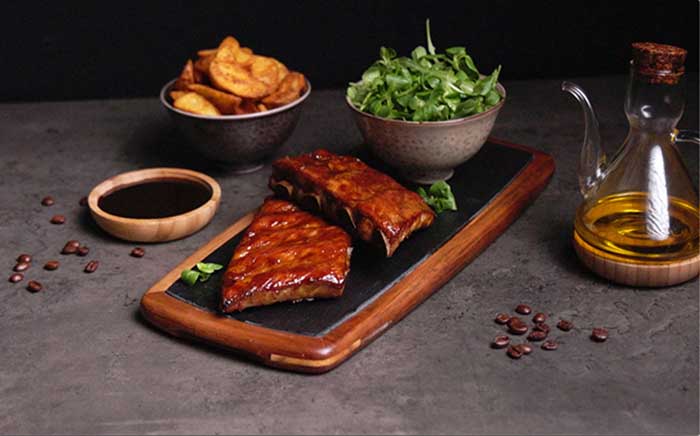 Costillas hechas en barbacoa de gas servidas en tabla de madera con pizarra con bol de canónigos y patatas al grill