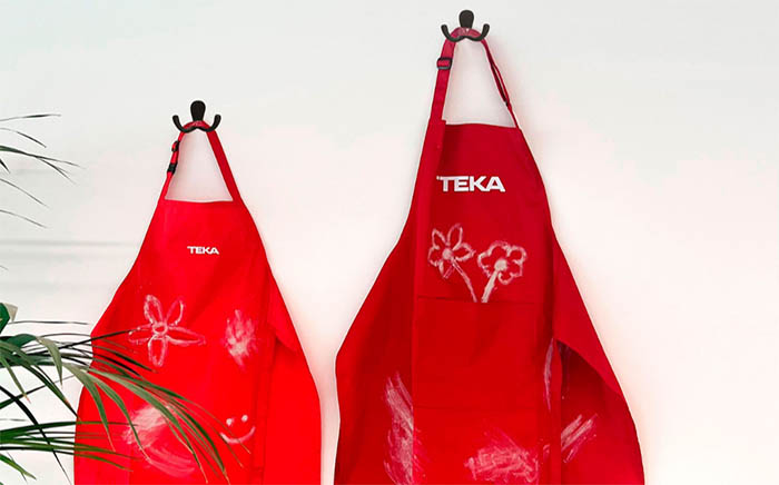 Pack delantales rojos Teka para adultos y niños colgados de un gancho en la pared blanca con manchas de harinas y dibujos