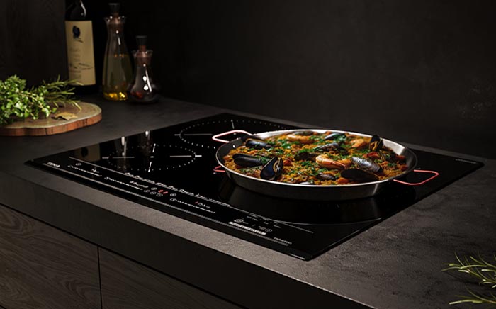 Placa Teka MestrePaeller sobre fondo oscuro y encimera negra, con paella recién hecha rodeada de romero e ingredientes para cocinar como aceite de oliva y perejil. Una opción para renovar tu cocina este Black Friday