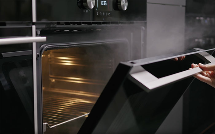 Teka's SteamGourmet oven with light inside and a hand opening the door while steam comes out from inside