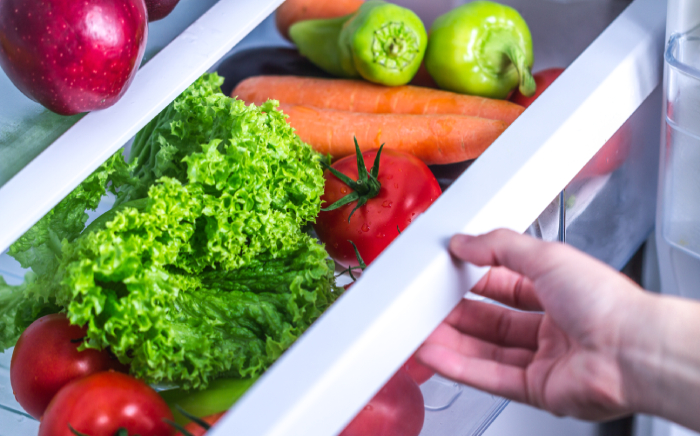 Mano abriendo un cajón del frigorífico con verduras frescas como zanahorias, tomates y pimientos como idea de cómo colocar la comida para ahorrar energía frigorífico