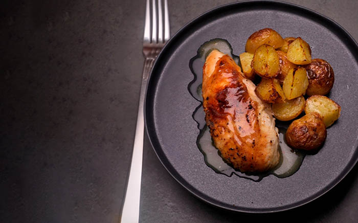 Plato negro de pollo son salsa asado en un horno con patatas y un tenedor a la izquierda sobre superficie negra