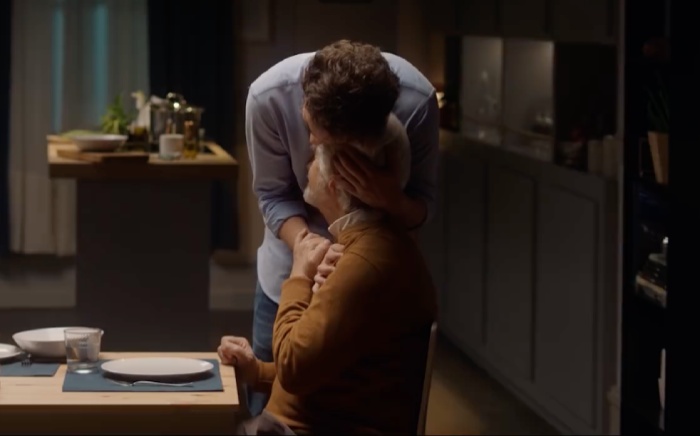 Un hijo adulto abraza a su padre adulto sentado a una mesa justo antes de sentarse a comer en familia en una imagen que liga comida con emociones