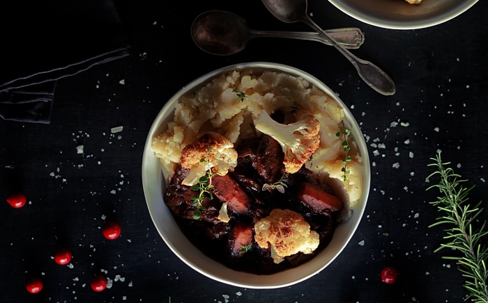 Cacerola con coliflor, heura y puré de patatas cocinados sobre una encimera negra con motivos navideños como ramitas de romer y frutas rojas junto a dos cucharas de plata