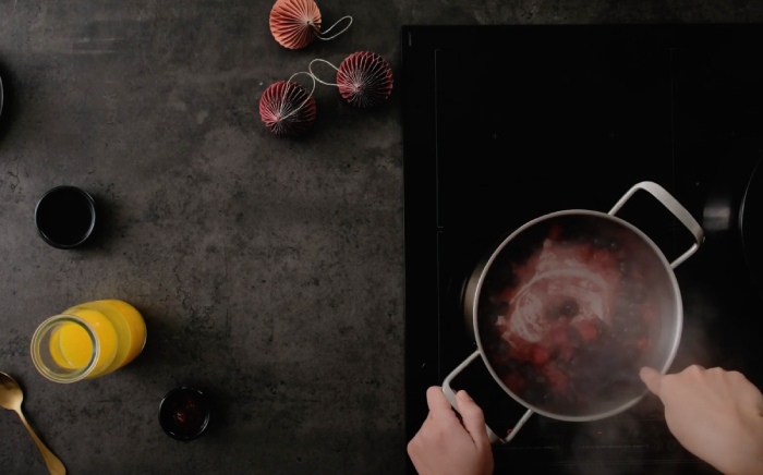 Plano cenital de una una encimera con motivos navideños y zumo de naranja y una placa donde unas manos cocinan en una cazuela frutos rojos con agua