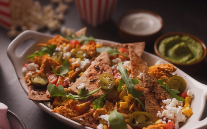 Fuente blanca de horno con nachos recién hechos con topping de queso cheddar, jalapeños, tomates cherry cortados y canónigos sobre una superficie con un cuenco de guacamole y otro de salsa agria