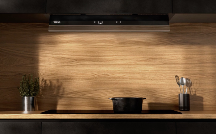 Vista frontral de una placa de cocina con una cacerola de color oscuro y una campana integrada Integra Pro de Teka de colore negro casi invisible con la visera desplegada en una cocina con la pared de madera y una planta artificial a la izquierda junto a utensilios de cocina a la derecha en un cubilete