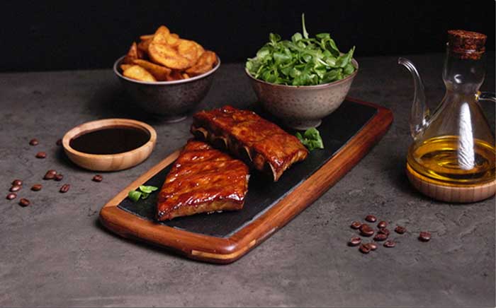 Ribs cooked on a barbecue served on a wooden cutting board with a bowl of rocket salad and grilled potatoes