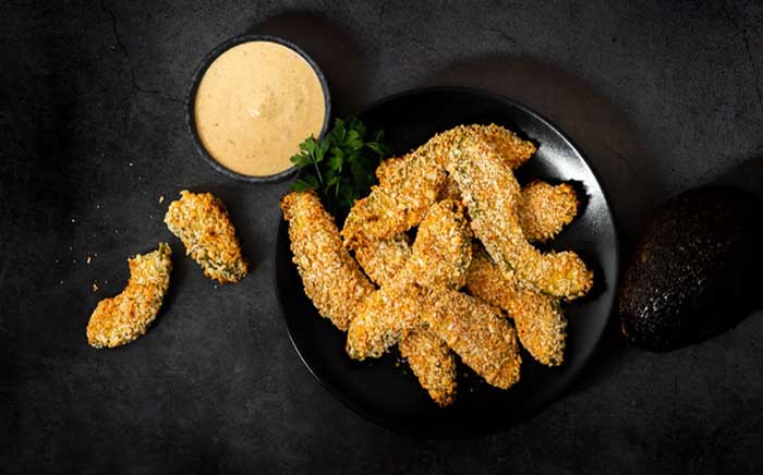 Fried avocado fingers on a black plate with a bowl of sauce