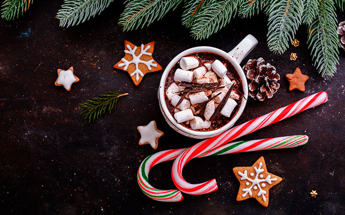 A cup of hot chocolate with marshmallows with Christmas sweets and decorations together with Christmas-themed cookies on a table