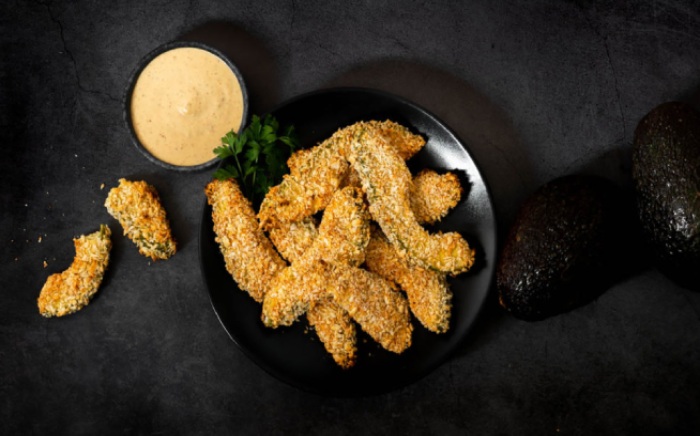 A bowl with crispy avocado fingers on a black surface together with some sauce to dip in