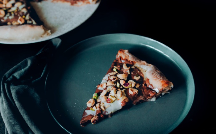 A blue green plate with a slice of chocholate pizza topping with nuts over a black surface 