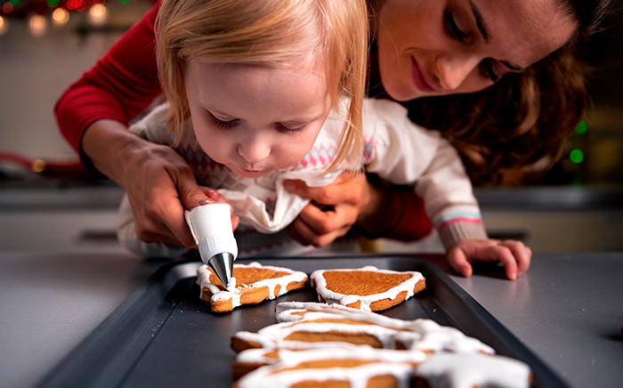 Woman holding a little girl while she is decorating cookies arranged on an oven tray ready for the oven