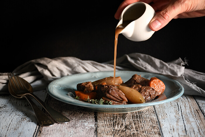 A hand pouring sauce over a plate of traditional meat stew with vegetables