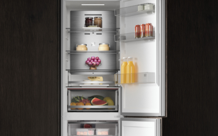 Close-up view of an open built-in refrigerator interior with stainless steel back panel, LED lighting, glass shelves, and organized food items.