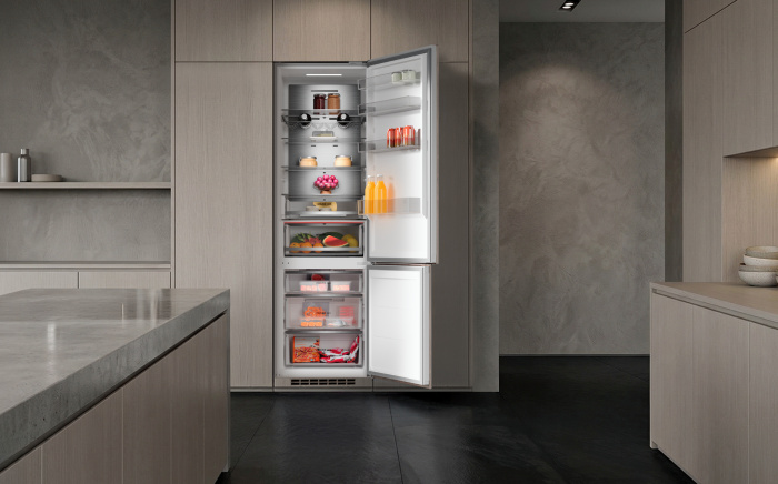 Open built-in fridge-freezer refrigerator stocked with fresh groceries and drinks, integrated into modern light wood cabinetry.