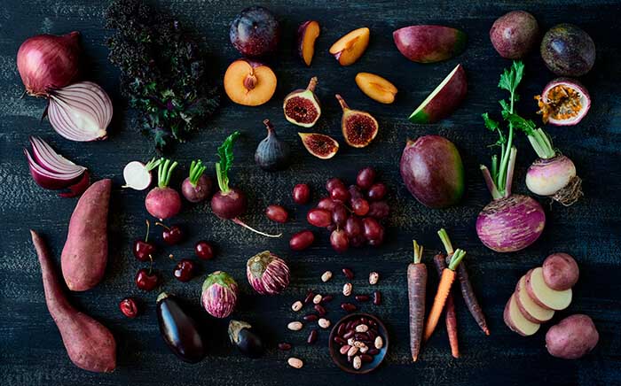 Flat lay composition of various raw purple and deep red fruits, vegetables, and legumes spread out on a dark background.