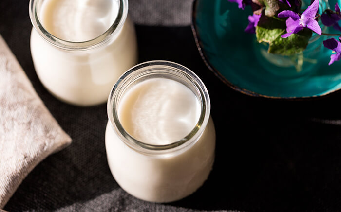 Two clear glass jars filled with creamy plain white yogurt on a dark cloth next to purple flowers