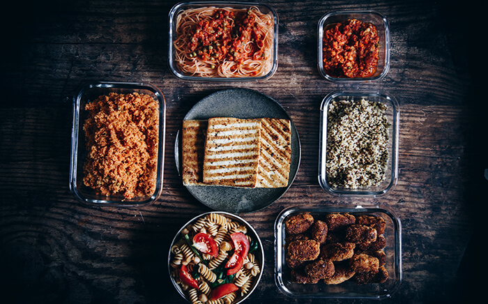 Flat lay view of meal prep containers for batch cooking and plates filled with cooked foods including grilled tofu, pasta, quinoa, seasoned rice, and nuggets on a dark wooden surface