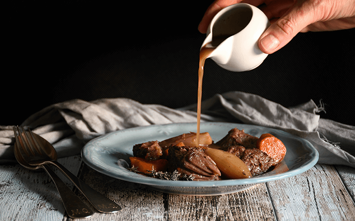 A white jar pouring sauce over a dish of meat stew with carrots, shallots, and rosemary