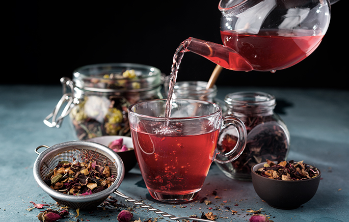 Infusión de color rojo servido en una jarra transparente y una taza de cristal rodeada de tés e infusiones