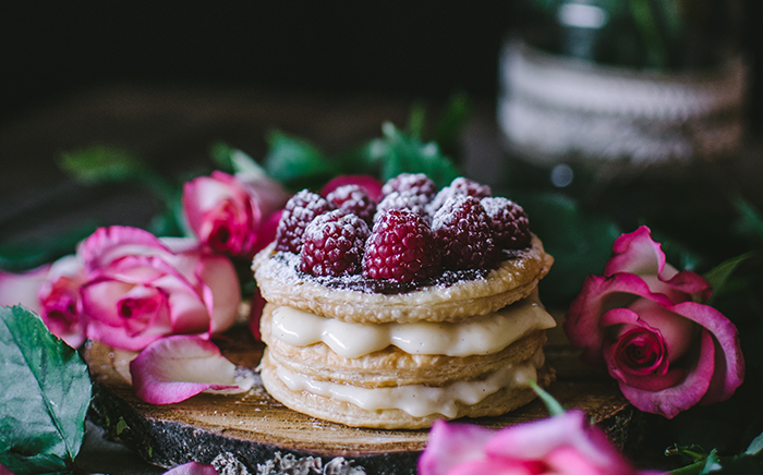 A pastry dessert with several layers and cream between them. The top has raspberries with icing sugar and it is surrounded by pink roses